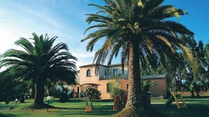 A building with a curved facade and a balcony, surrounded by palm trees and a well-maintained lawn. - Studio with air conditioning (Pula Sardinien)