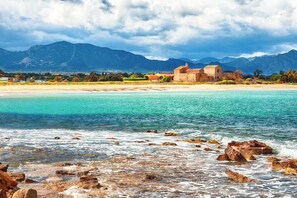 A coastal scene with a beach, clear turquoise waters, rocky shoreline, and a building with a red roof in the background. - Studio with air conditioning (Pula Sardinien)