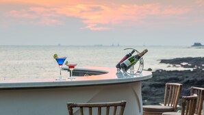 Un bar proposant deux cocktails et une bouteille de champagne, avec vue sur une côte rocheuse et un magnifique coucher de soleil.