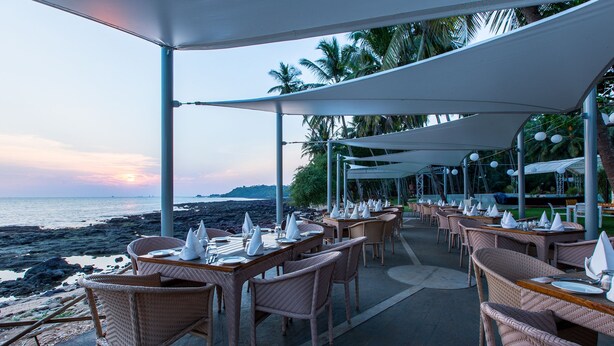 Un restaurant en bord de mer, avec des chaises et des tables en osier, des auvents blancs et une vue sur l’océan et la plage au coucher du soleil.