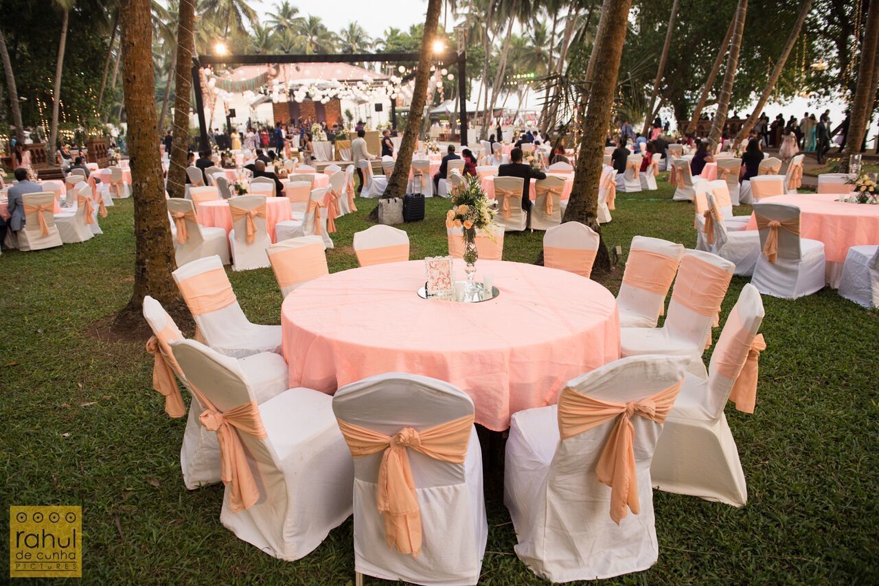 Una recepción de boda al aire libre con mesas redondas cubiertas con manteles rosas y sillas blancas con cintas color melocotón. Invitados sentados alrededor de las mesas, y un escenario montado al fondo.