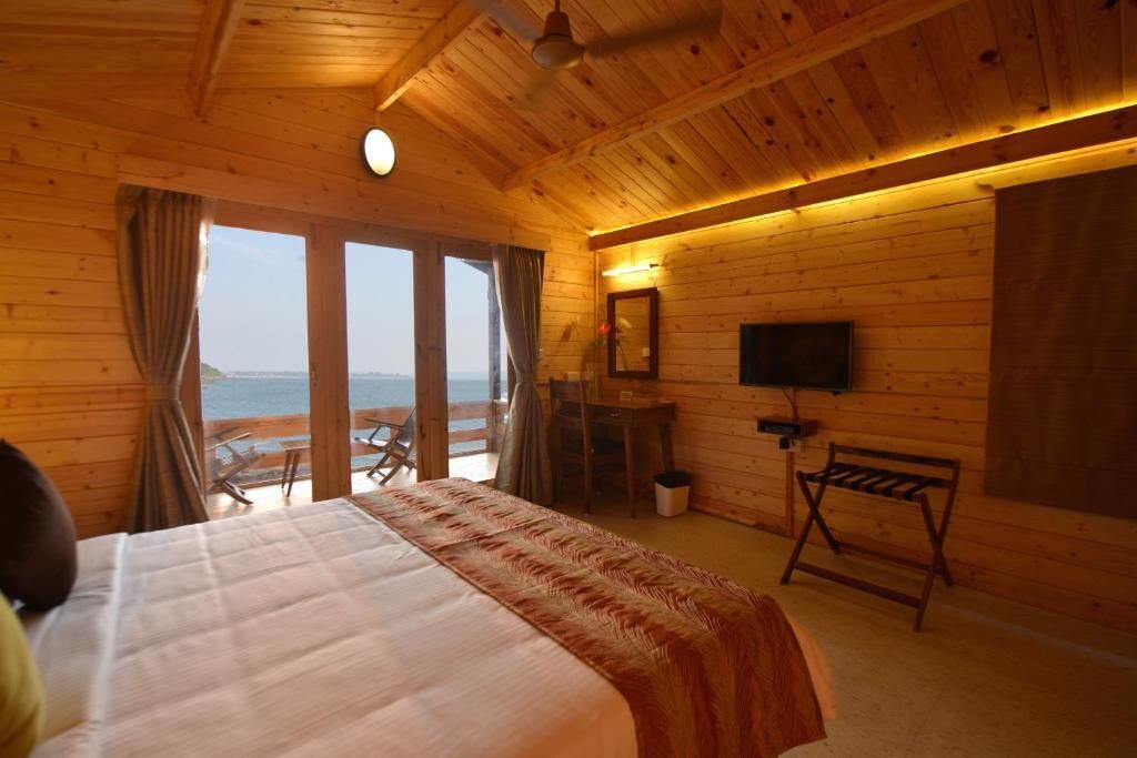 A wooden cabin room with a bed, a desk, a chair, a TV, and a view of the ocean.