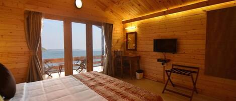 A wooden cabin room with a bed, a desk, a chair, a TV, and a view of the ocean.