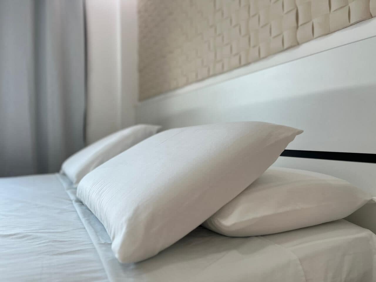 Una cama bien hecha con ropa blanca y almohadas, un listón negro en la cabecera y una pared texturizada al fondo.