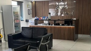 A hotel reception area with a wooden reception desk, two black chairs, a refrigerator, and a tiled floor. - Goiana Palace Hotel (Goiana)