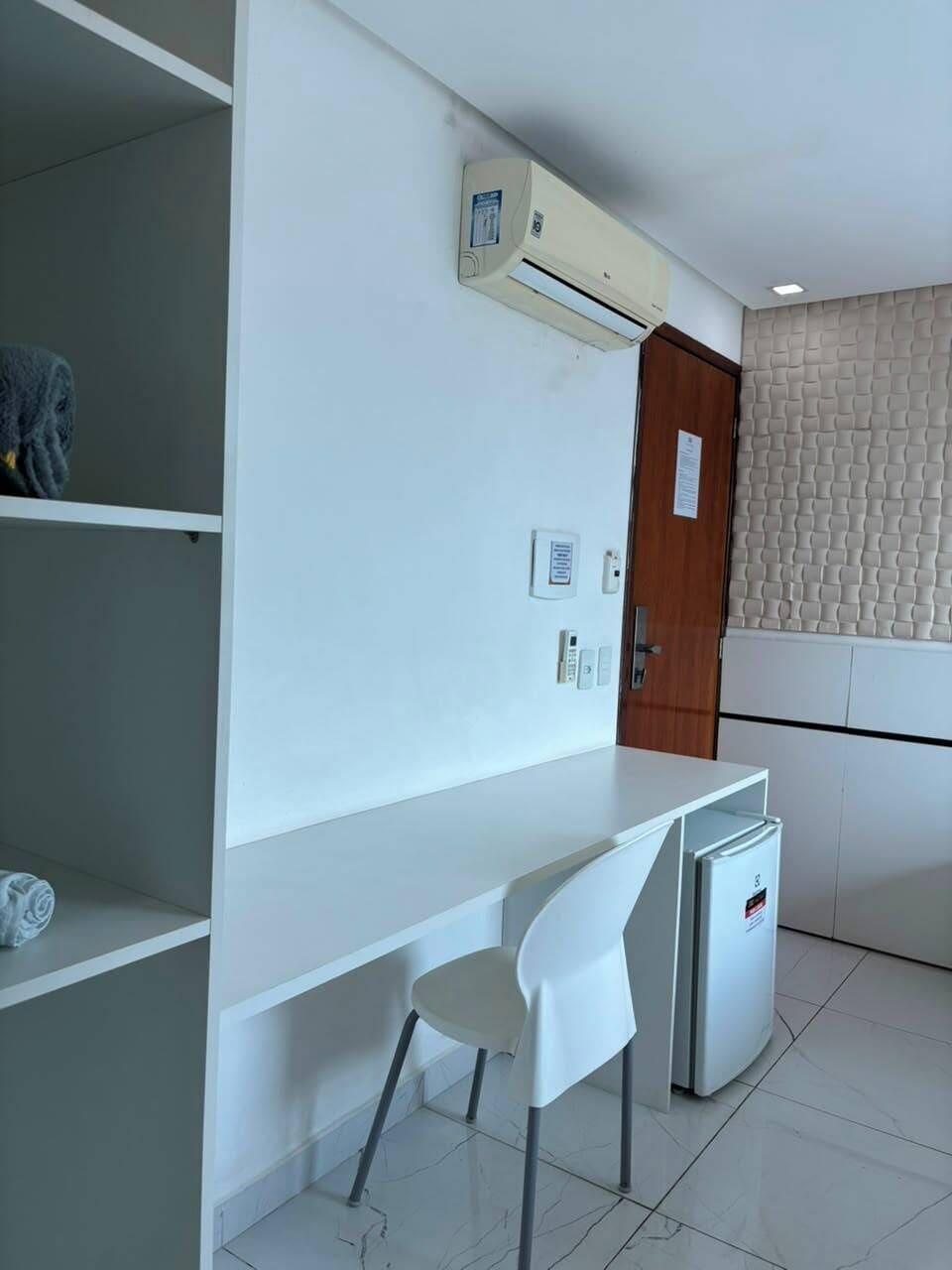 A room with a white desk, a chair, a small refrigerator, an air conditioning unit, a door, and a textured wall.