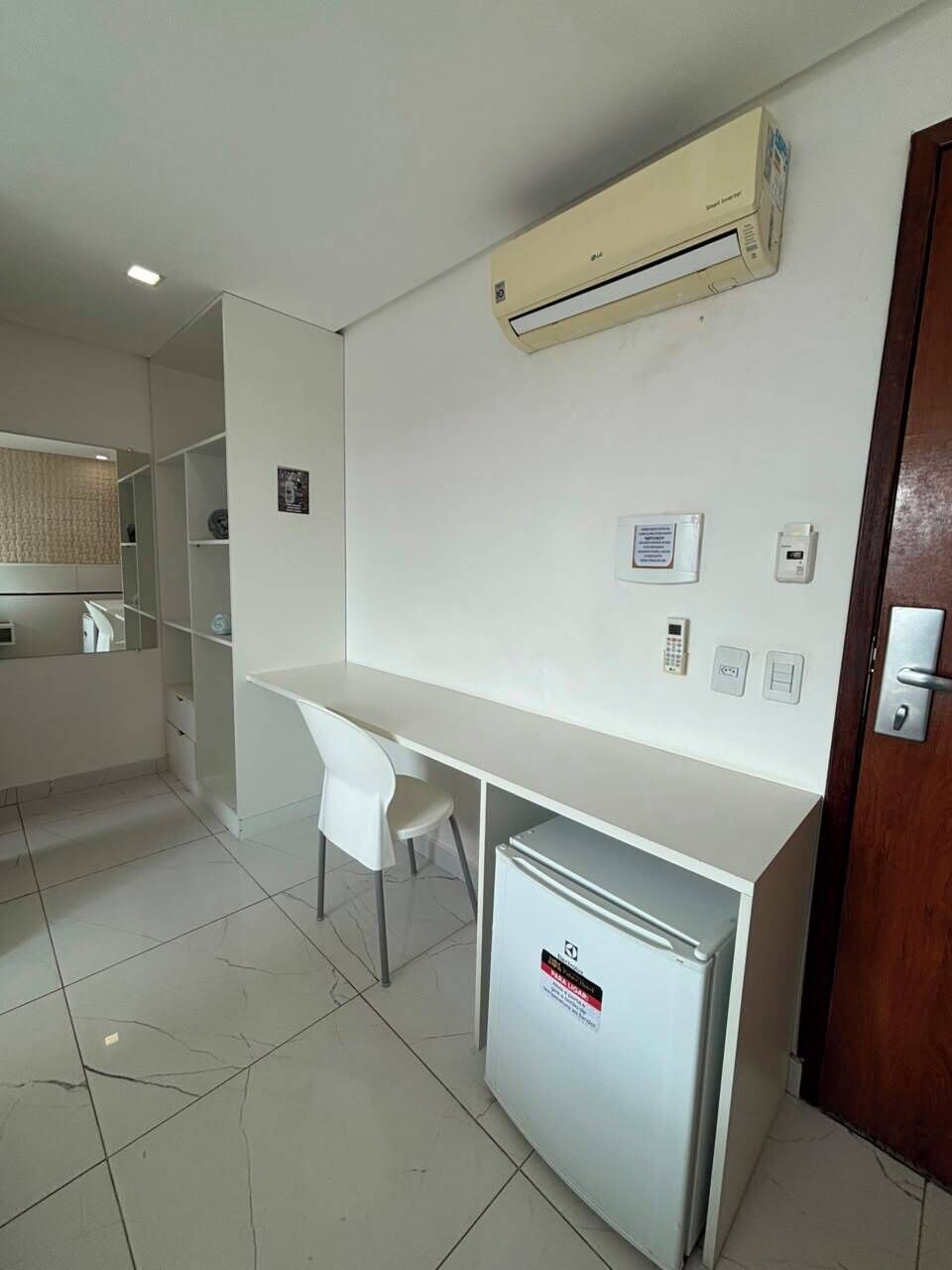 A hotel room with a white desk, a small refrigerator, an air conditioning unit, and a door with a lock.