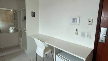 Ett hotellrum med ett vitt skrivbord, ett litet kylskåp, en luftkonditioneringsanläggning och en dörr med ett lås.