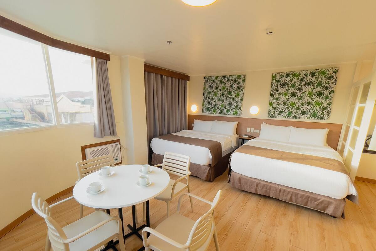 A hotel room with two beds, a dining table with chairs, and a view of the city.