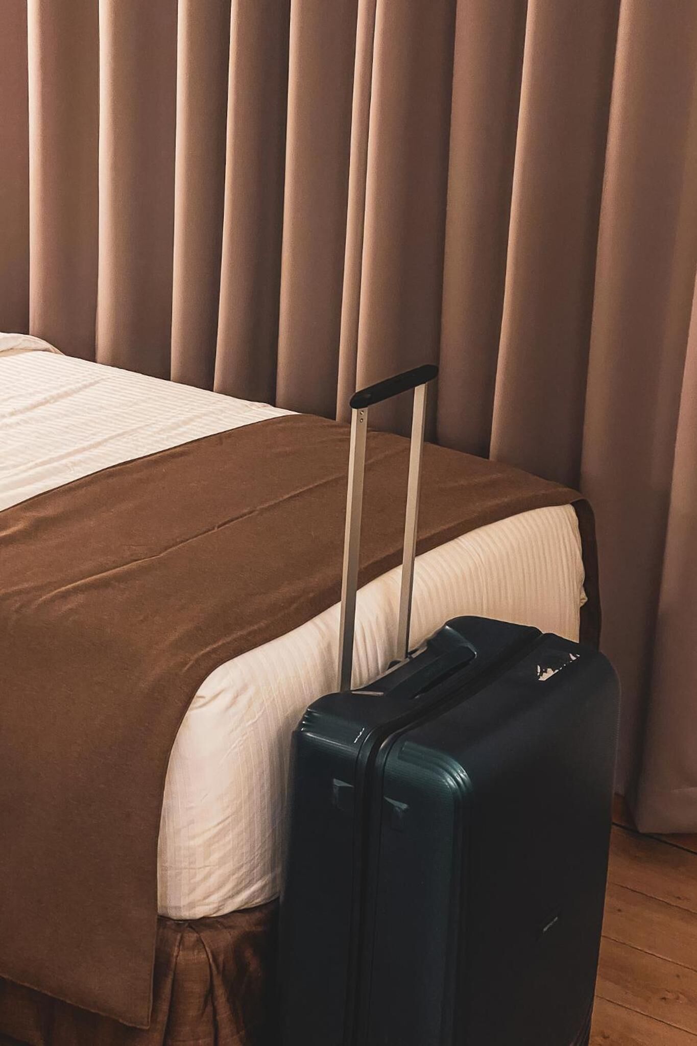 A bed with a brown bedspread and a blue suitcase with wheels beside it.