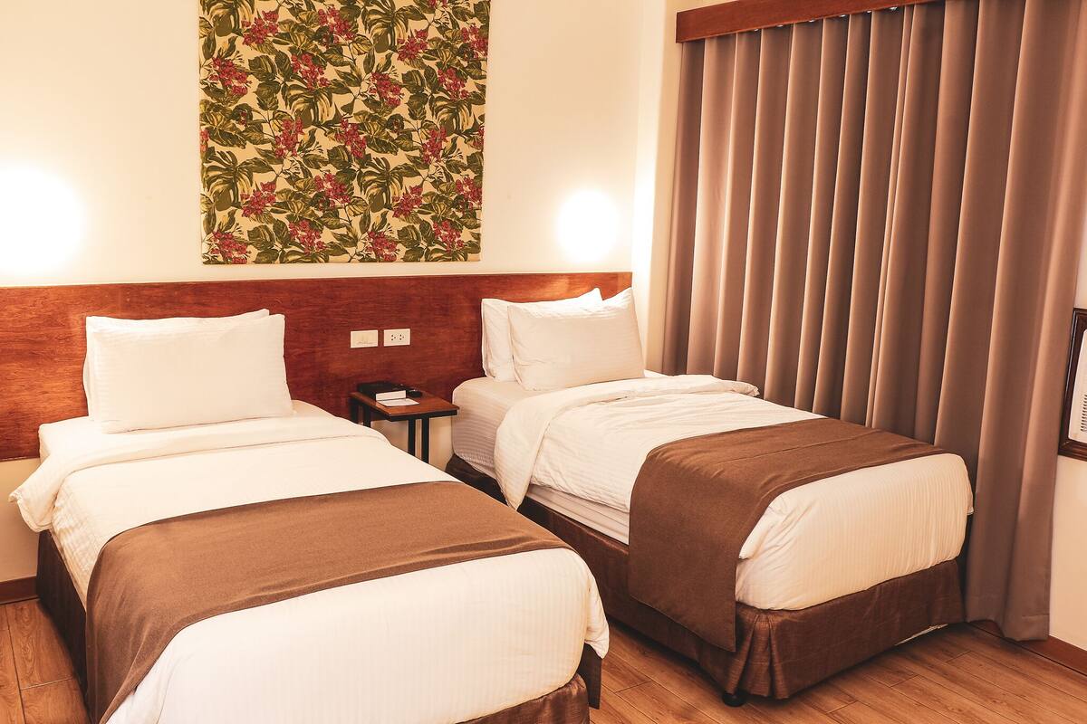 A hotel room with two beds, a wooden headboard, a nightstand, and a floral wall hanging.
