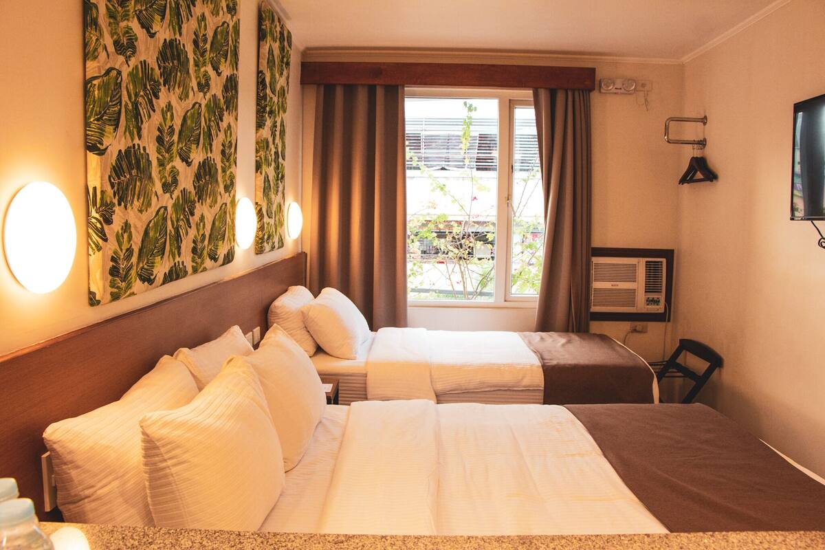 A hotel room with two beds, a window with curtains, a wall-mounted TV, and a wall-mounted heater.