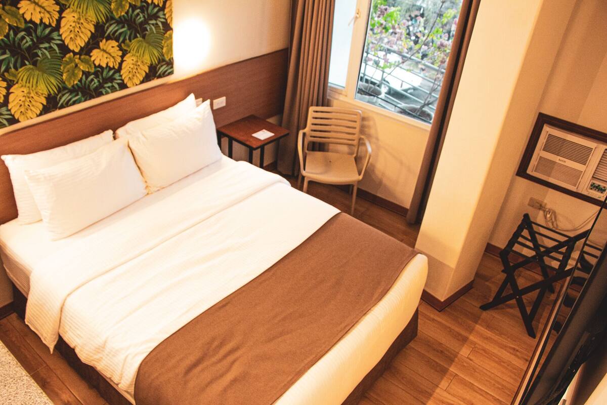 A hotel room with a large bed, a small table, a chair, and a window with curtains.