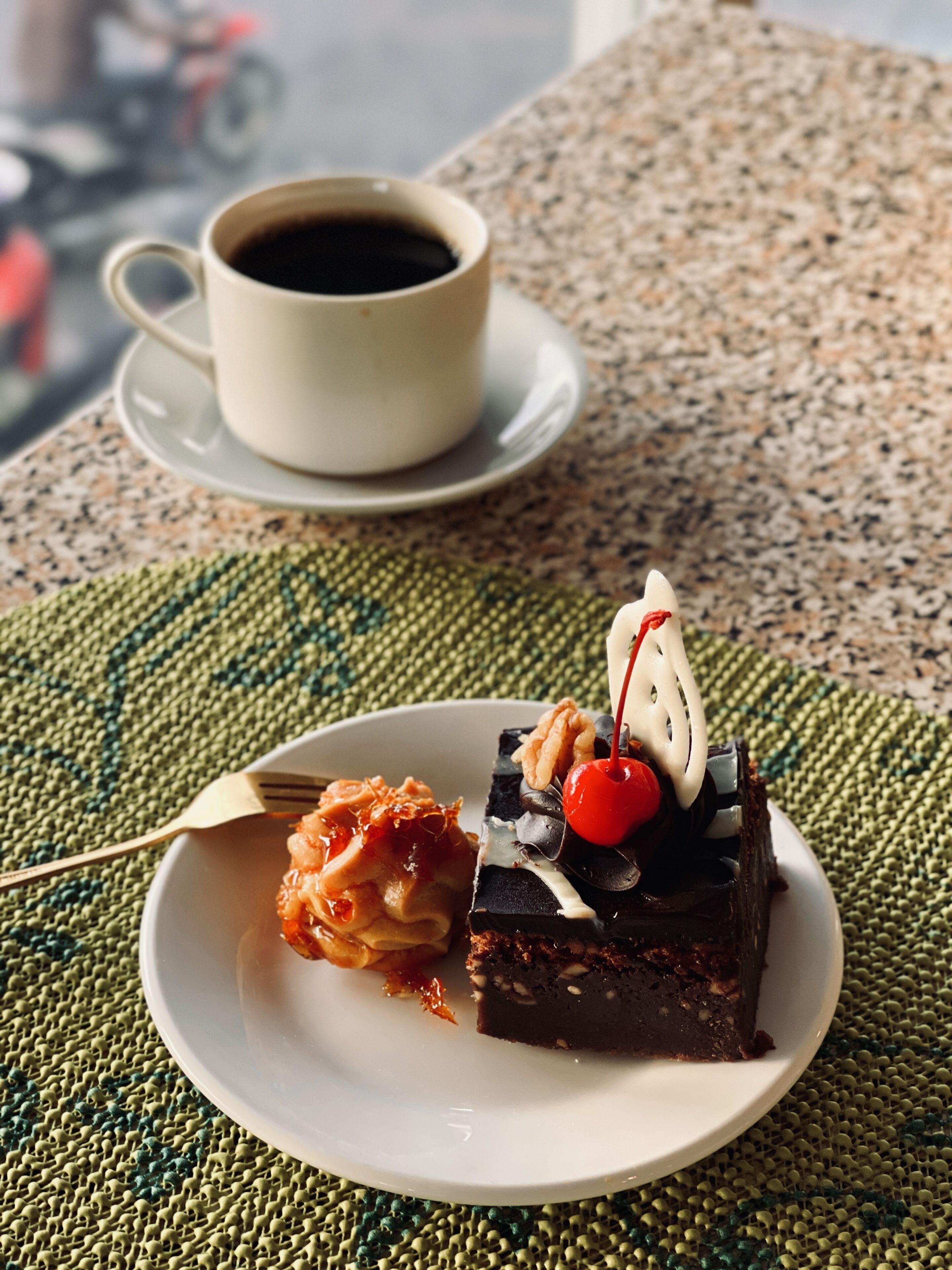 A dessert plate with a slice of cake and a cherry on top, a cup of coffee, and a fork on a woven placemat.