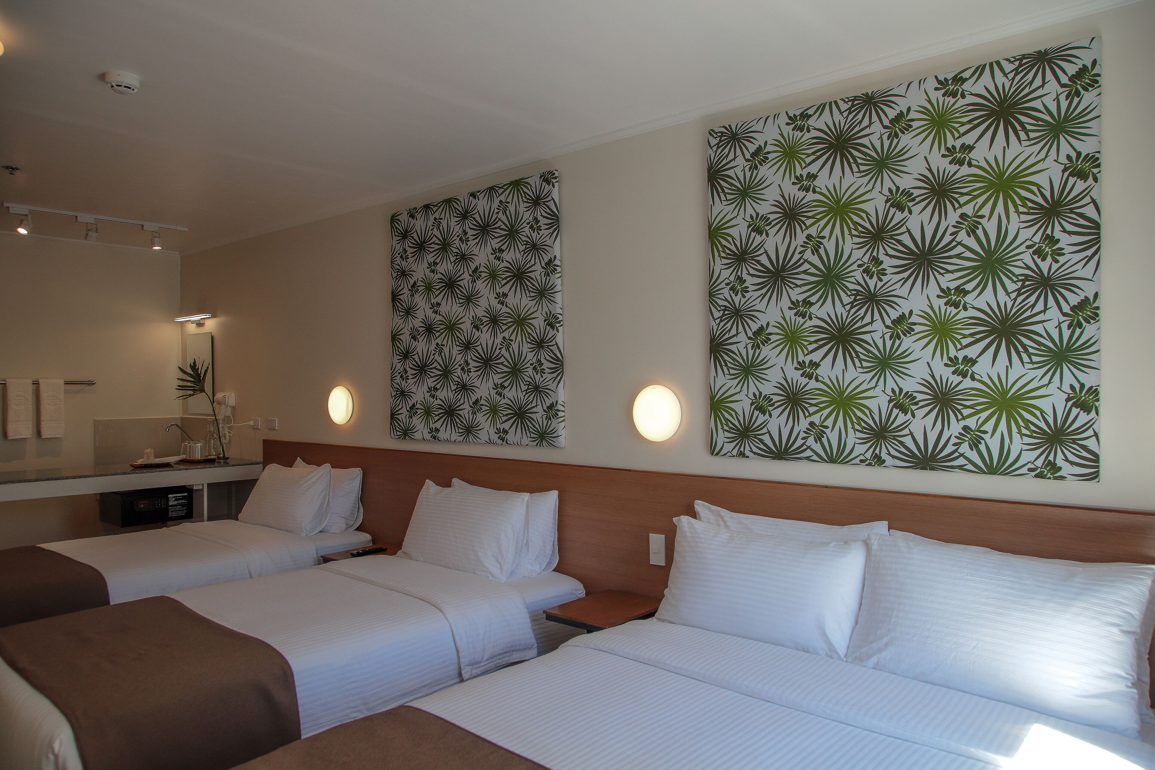 Habitación de hotel con dos camas, cabecera de madera, mesita de noche, espejo y cuadro con un motivo de hojas verdes.