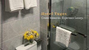 A modern bathroom with a glass shower enclosure, a toilet, and a sink. There are towels hanging on a rack and a vase with flowers on the sink. - Hotel Tugos (Baguio)
