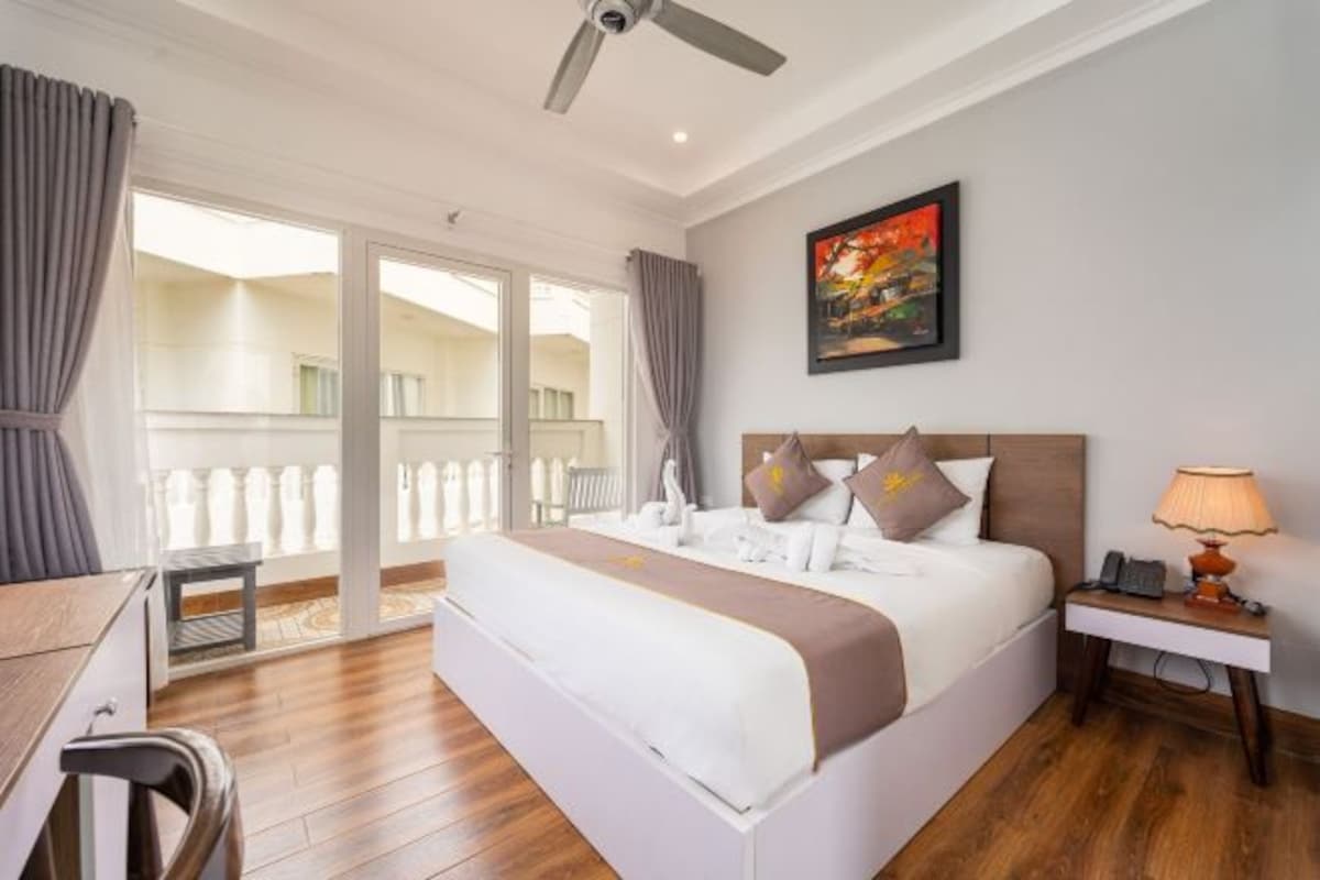 Ein Hotelzimmer mit Bett, Schreibtisch, Stuhl, Nachttisch mit Lampe, einem Bild an der Wand und einem Balkon mit Vorhängen.