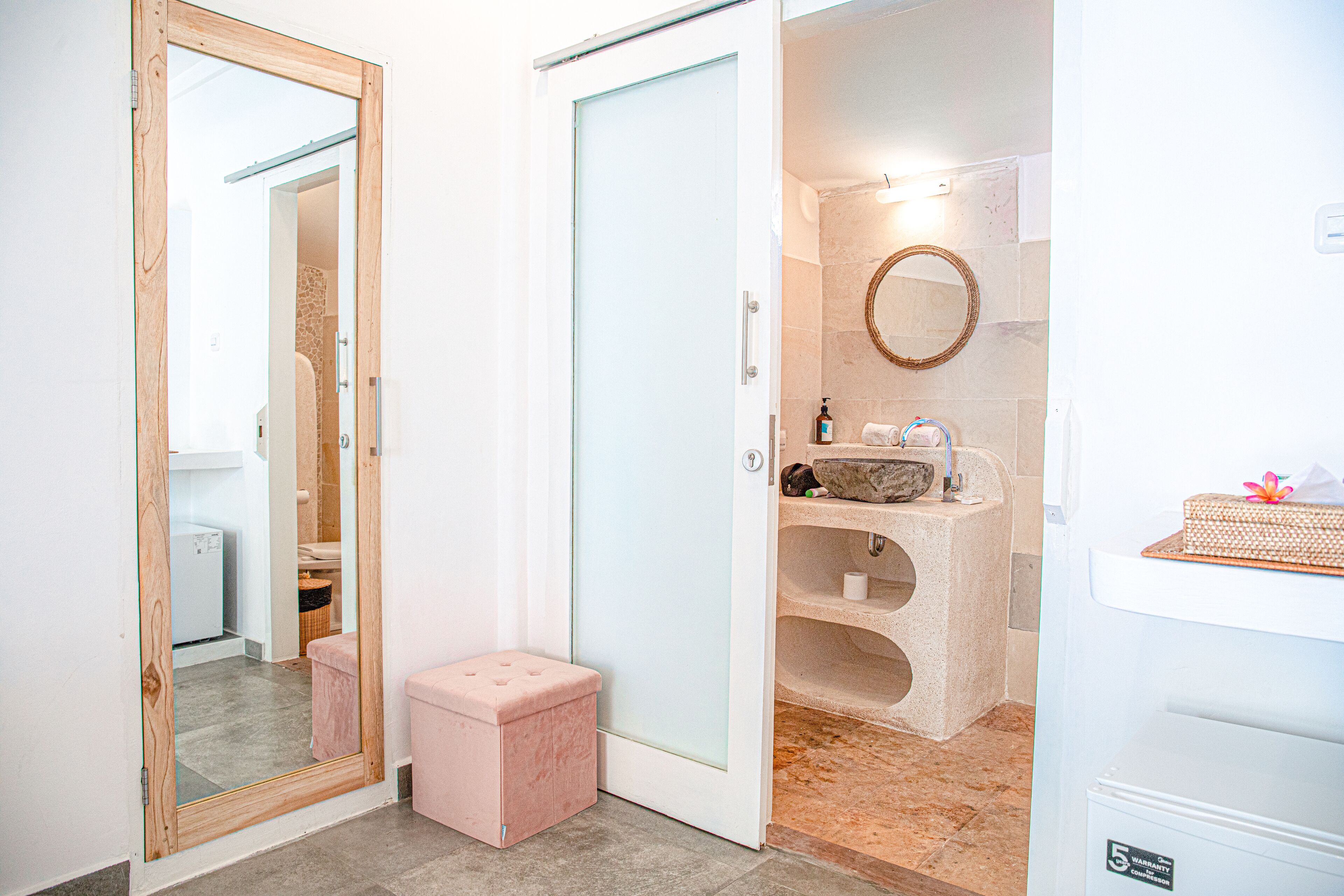A bathroom with a glass door, a wooden-framed mirror, a pink stool, a marble floor, and a sink with a towel and soap dispenser.