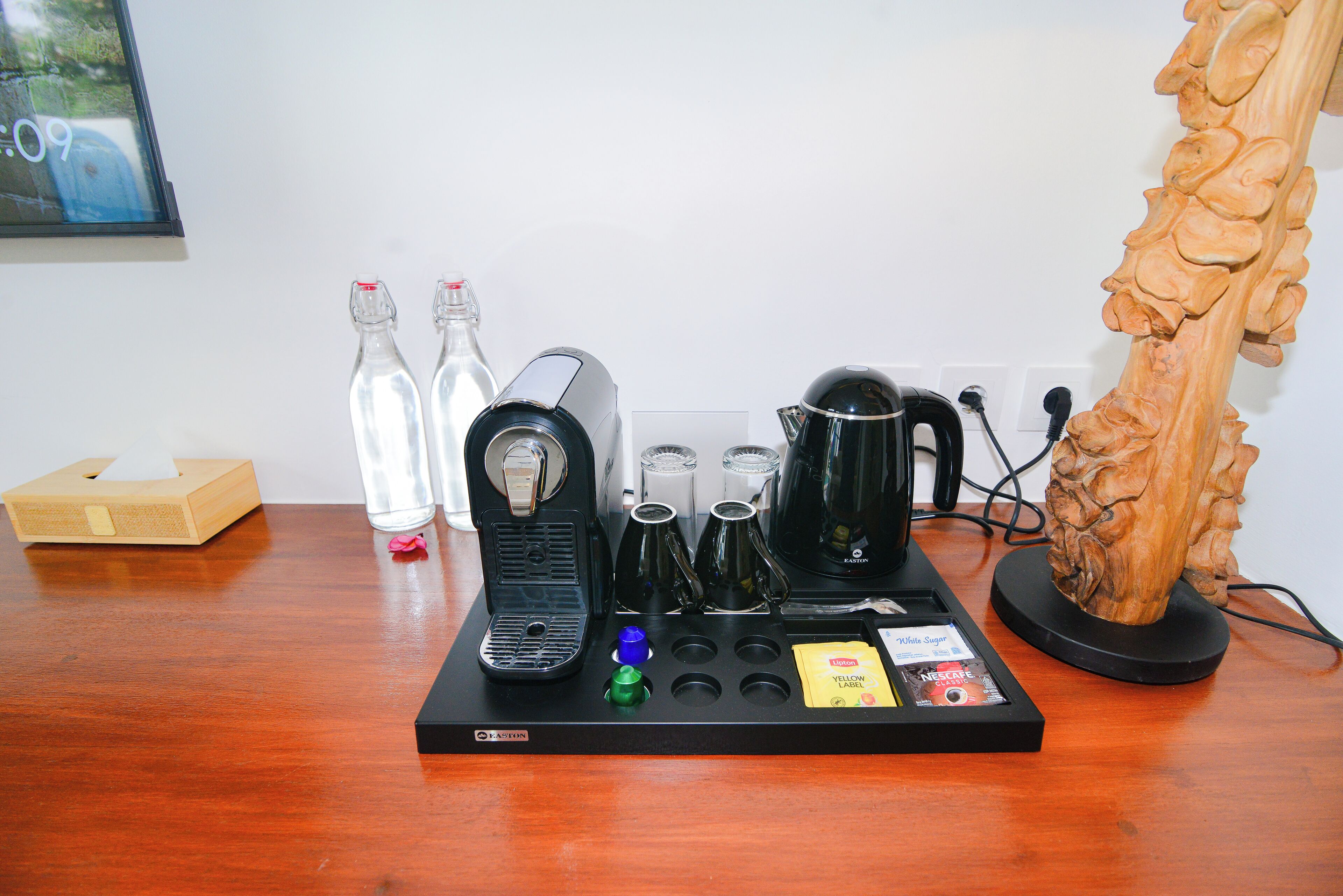 A coffee machine setup with various coffee pods, a tissue box, and a decorative wooden sculpture on a wooden table.