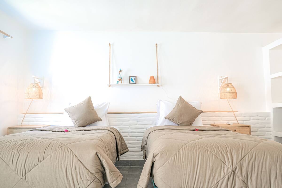 Une chambre d’hôtel avec deux lits, un mur blanc, une petite étagère avec des objets décoratifs et des lampes de chevet.