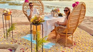 Een eettafel aan het strand met rieten stoelen, bloemen en een drankje.