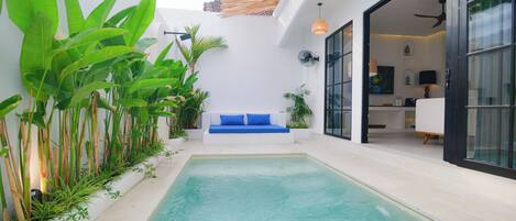 Un área de piscina moderna al aire libre con un sofá azul, rodeada de abundante vegetación y un muro blanco.
