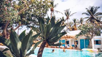 A pool area with palm trees, a building with blue doors, and people swimming.
