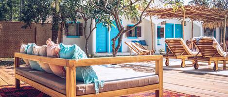 A wooden bed with pillows and a blanket, a wooden bench, and a patterned rug on a wooden deck.