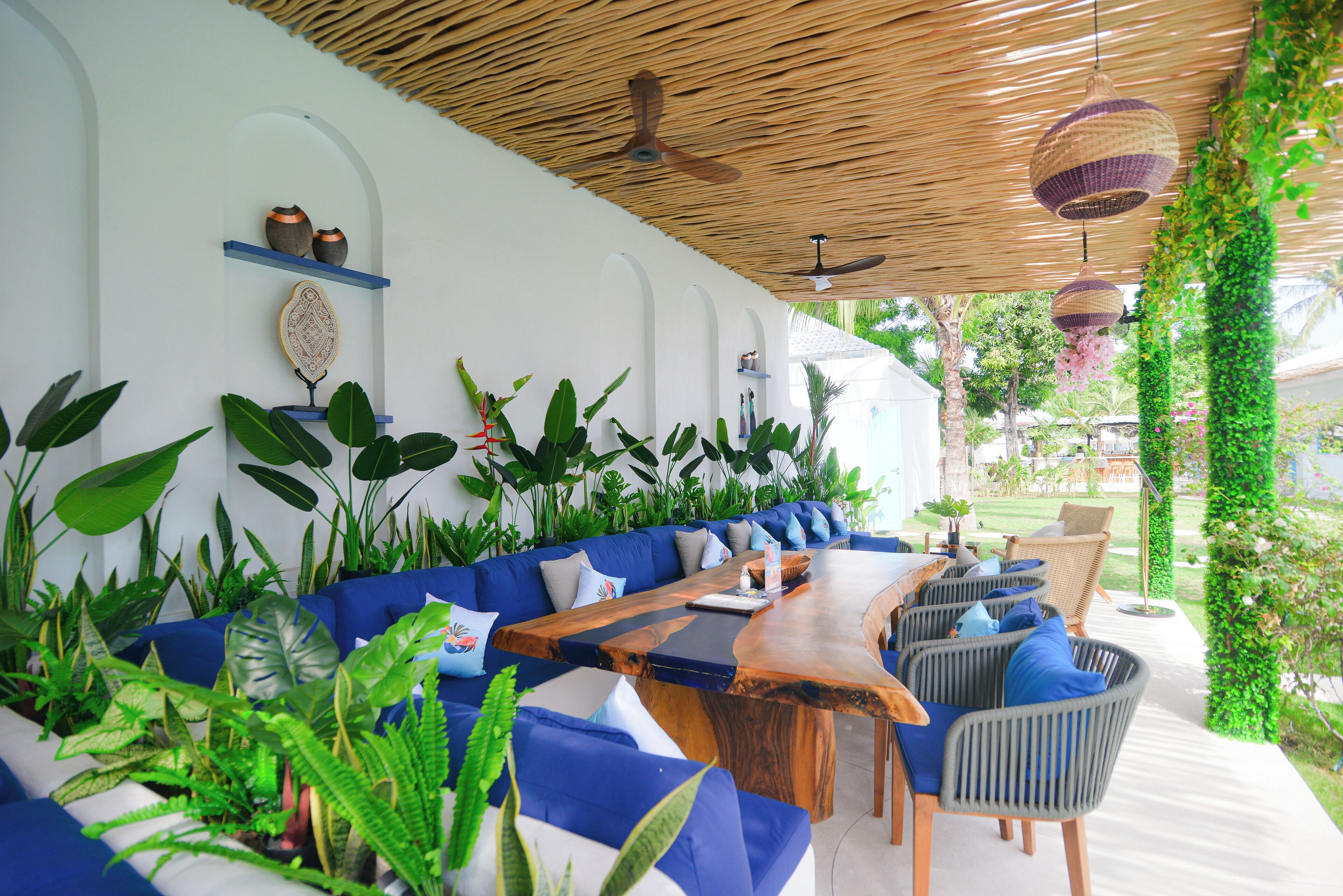 A modern outdoor seating area with a wooden table, blue cushioned sofas, and various plants.