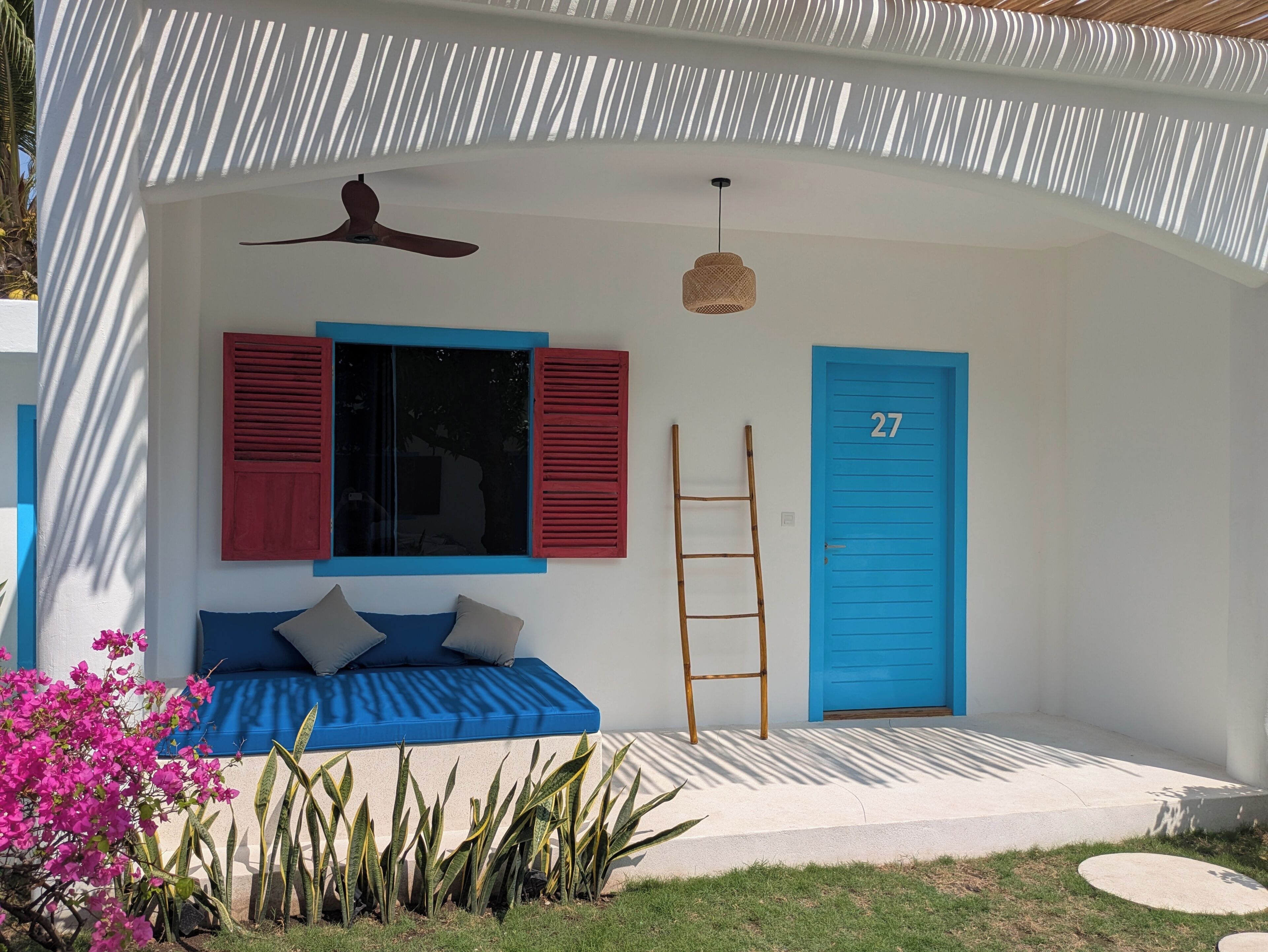 A modern house with a blue door marked with the number 27, a blue window with red shutters, a white wall, and a blue bench outside.