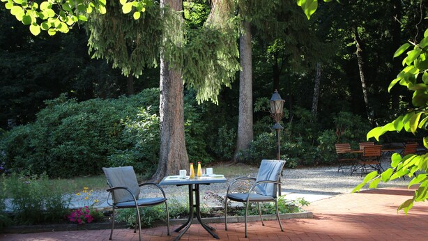 An outdoor patio with a table and chairs, surrounded by greenery and trees.