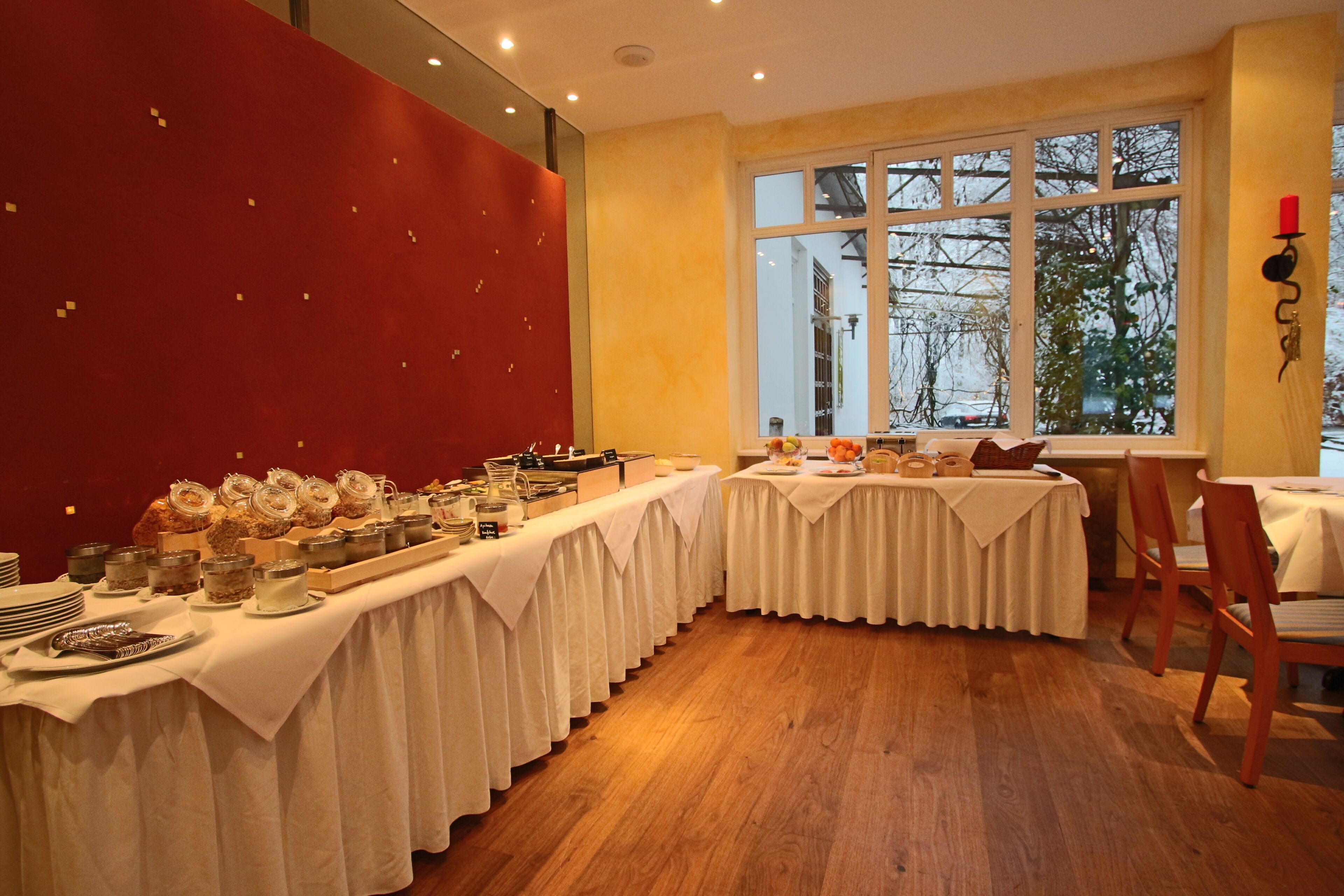 A dining area with long tables covered in white tablecloths, wooden flooring, and a large window with a view of trees.