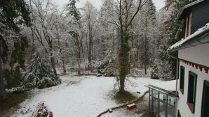 A snow-covered landscape with a house featuring a balcony and a window with a view of the snowy forest.