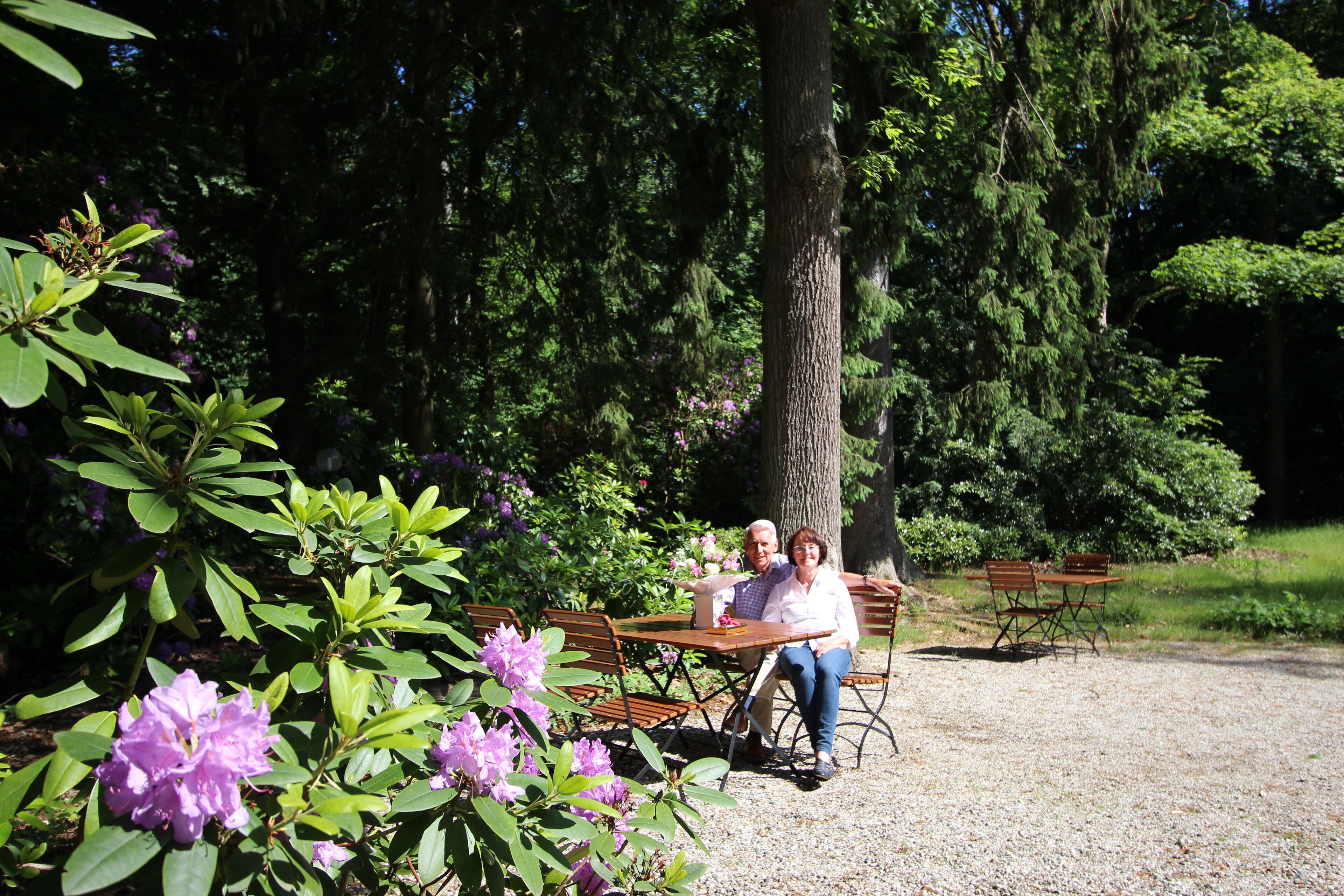 Un couple est assis à une table en bois dans un jardin agrémenté de bancs et d’arbres.