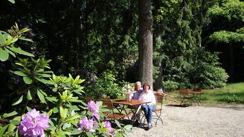 Un couple est assis à une table en bois dans un jardin agrémenté de bancs et d’arbres.