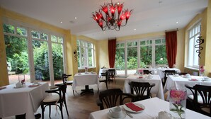 A dining area with multiple tables and chairs, large windows with curtains, and a chandelier.