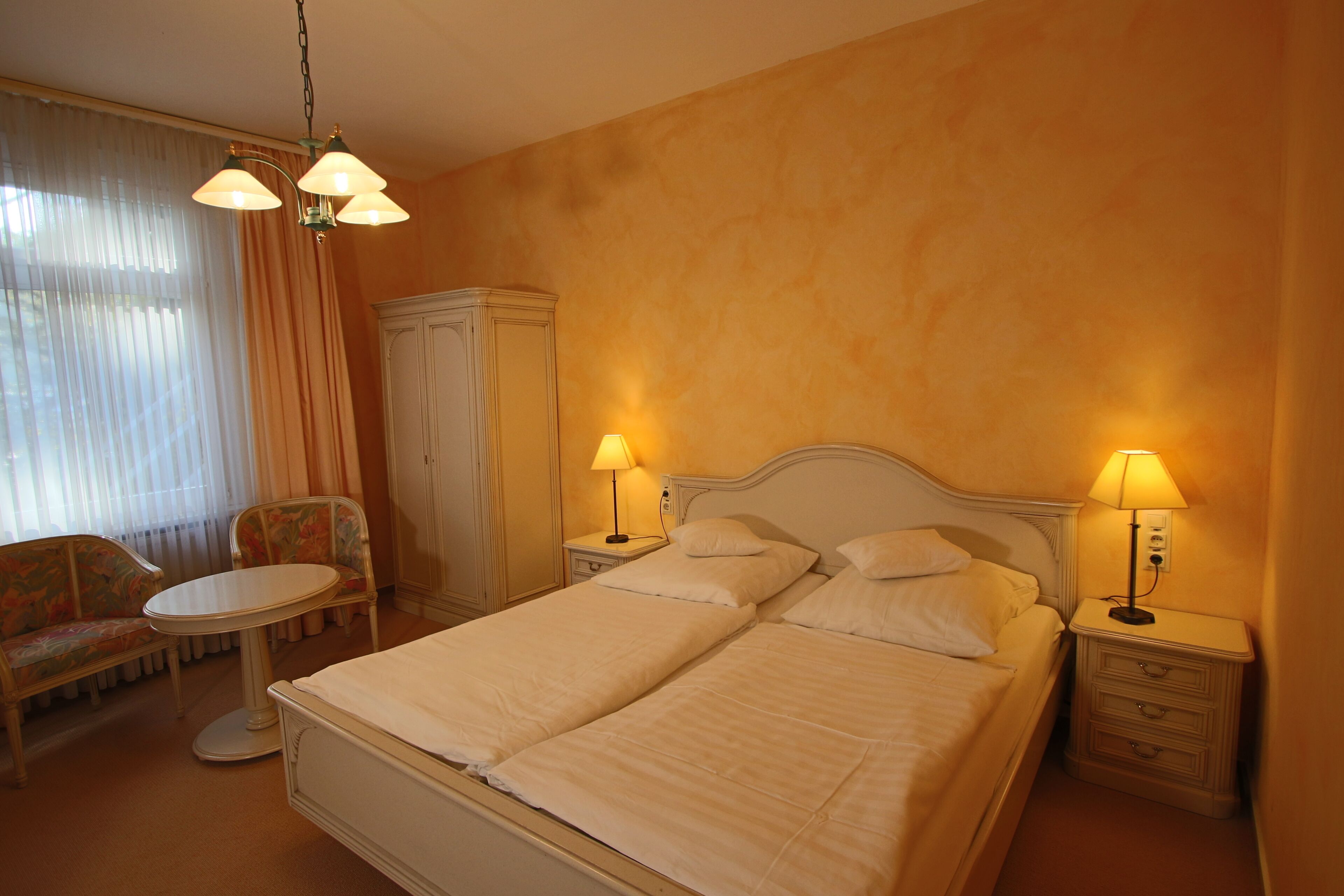 A hotel room with a double bed, two bedside tables with lamps, a chair, a small table, and a wardrobe.