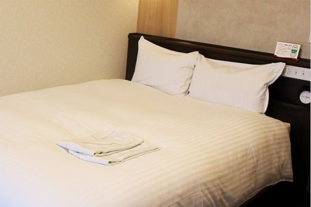 A neatly made bed with white linens and pillows, a wooden headboard, and a wall-mounted thermostat.