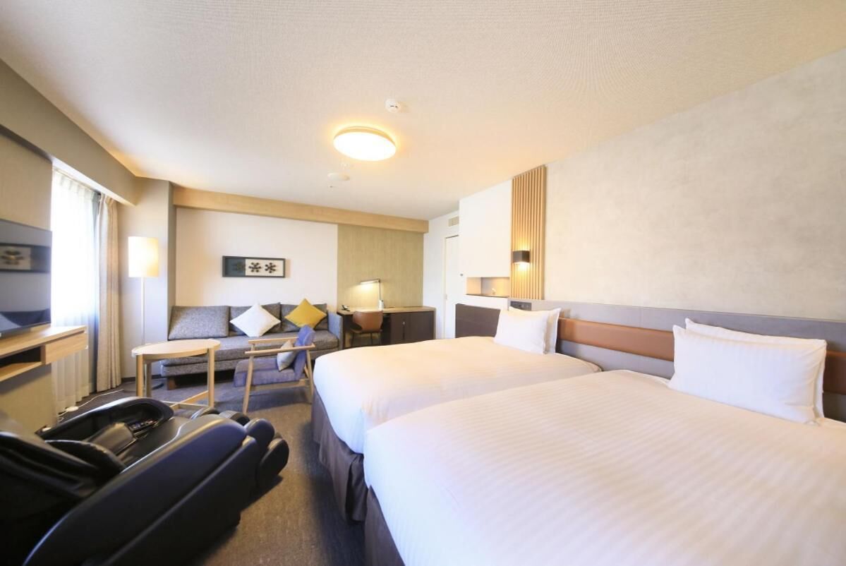 A hotel room with two beds, a desk, a chair, a sofa, and a TV.