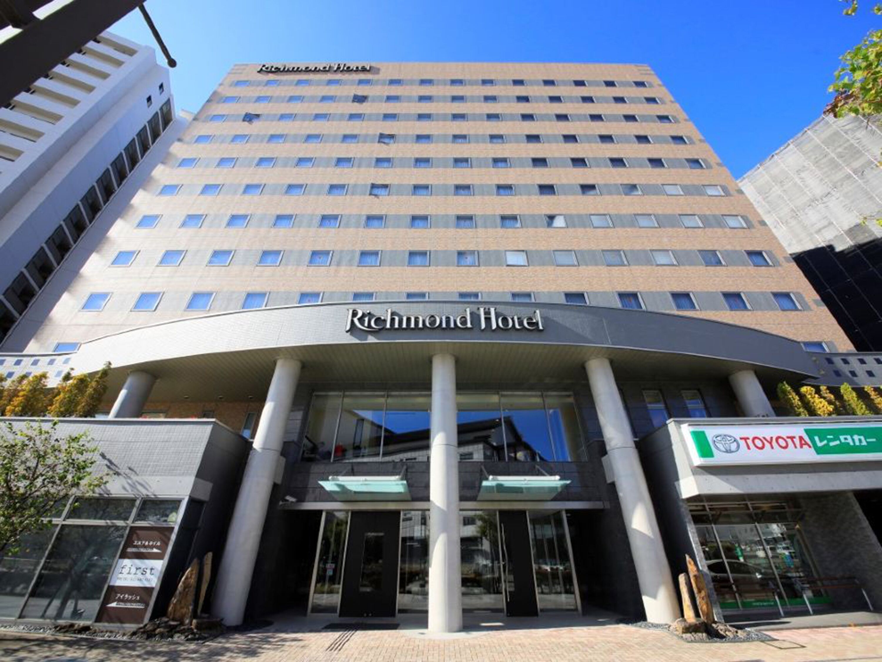 Un edificio de hotel alto de diseño moderno, con una entrada destacada y un cartel visible de una concesionaria Toyota.