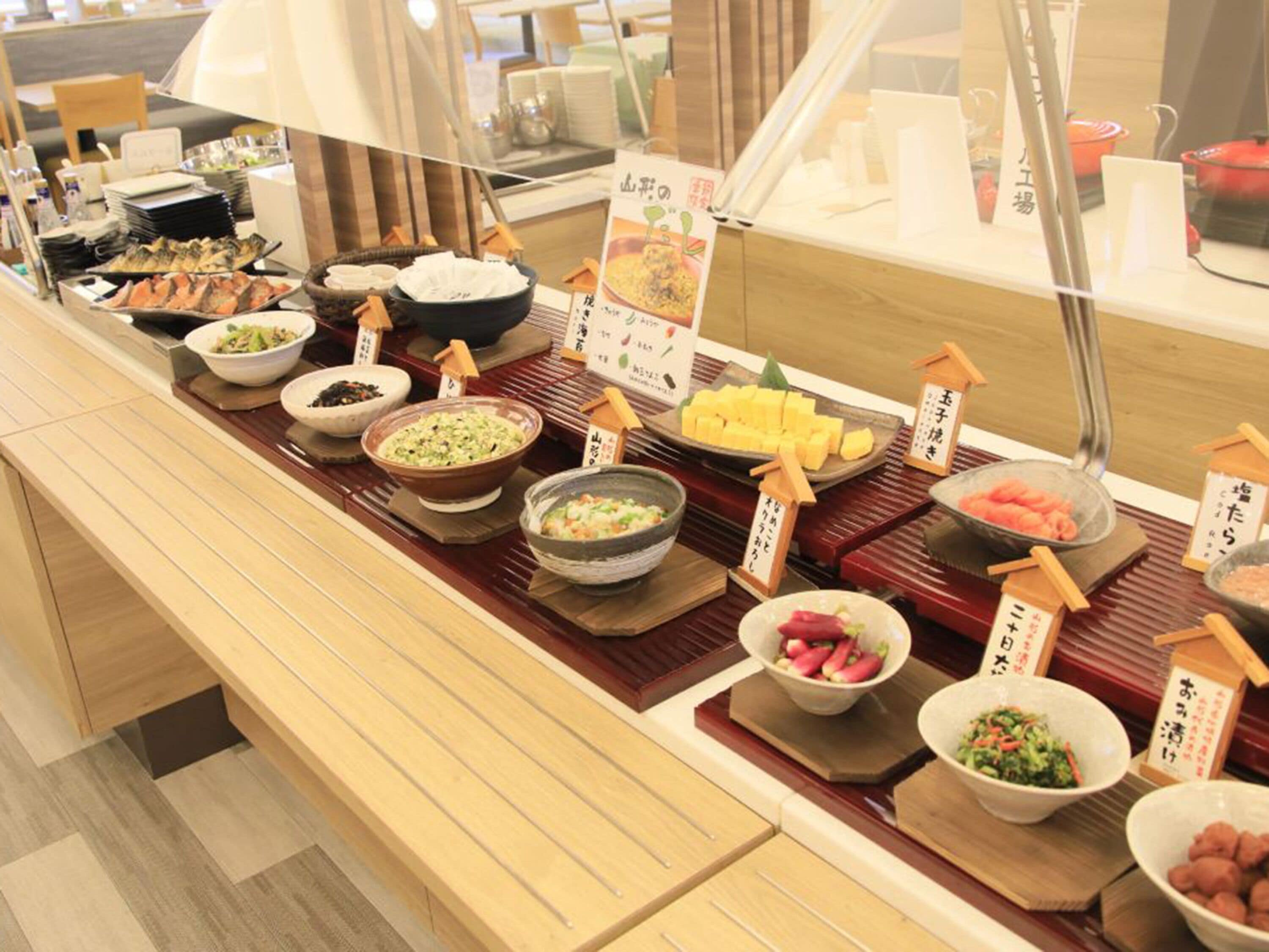 Un mostrador de buffet con diversos platos expuestos en cuencos y fuentes, cada uno con texto en japonés.