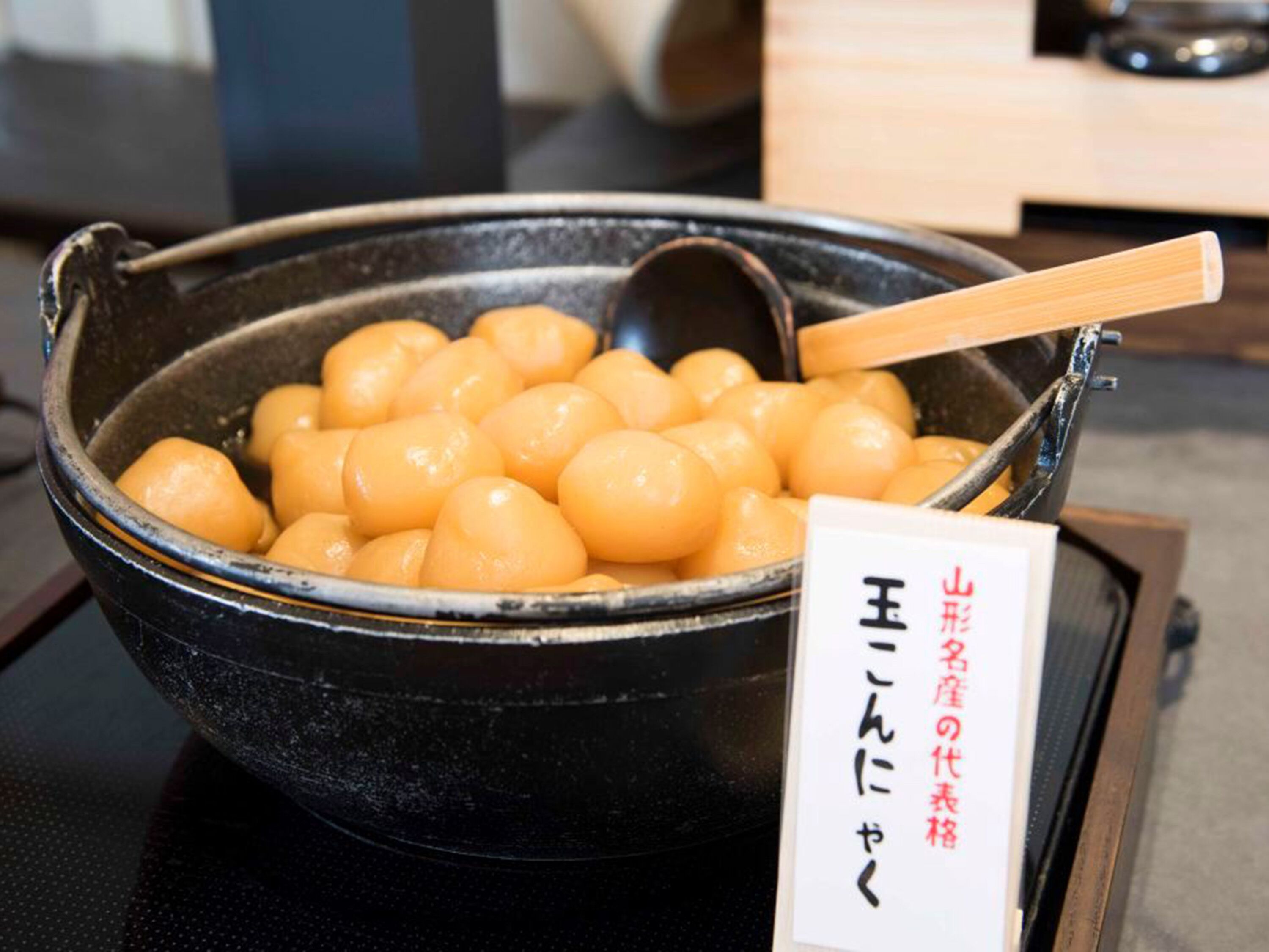 Una pentola nera piena di gnocchi gialli e rotondi, con un cucchiaio di legno all'interno. Accanto alla pentola, un cartello con scritte giapponesi.