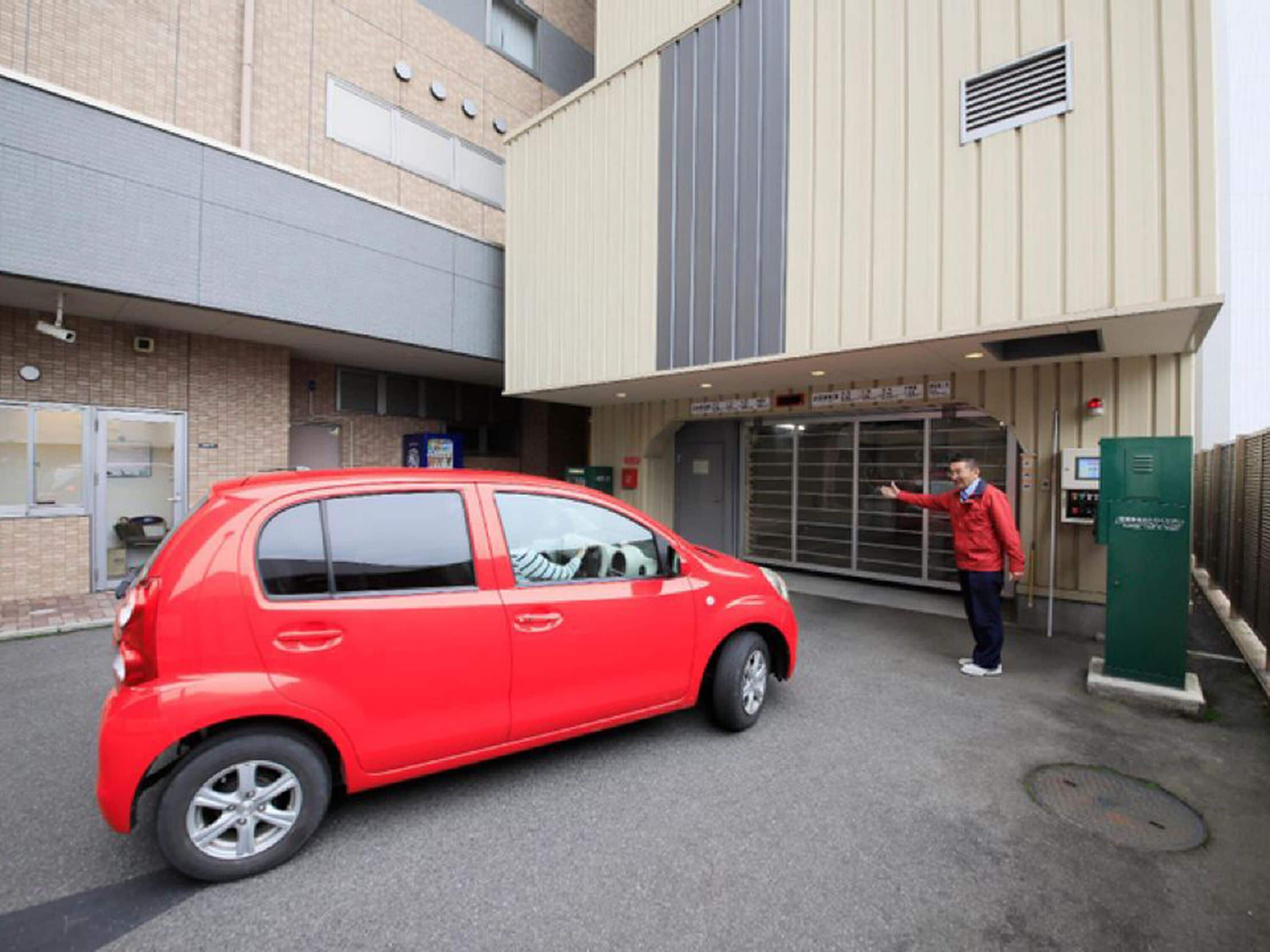 A red car parked in front of a building with a large entrance and a man gesturing towards it.