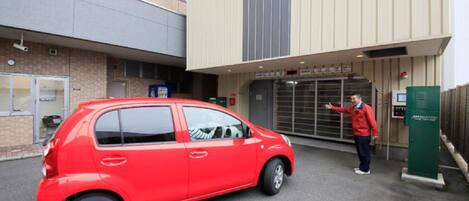 A red car parked in front of a building with a large entrance and a man gesturing towards it.