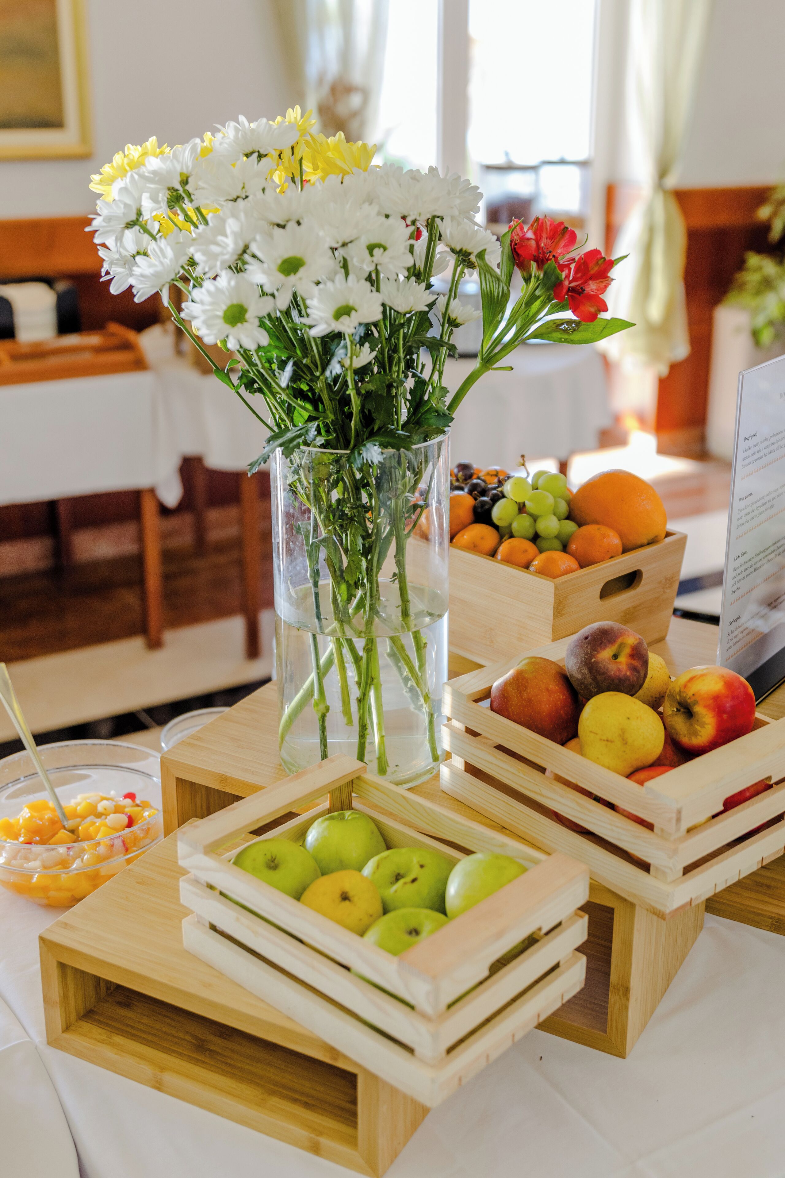 Una mesa con un florero de flores, cajas de madera con frutas y un tazón de ensalada de frutas.