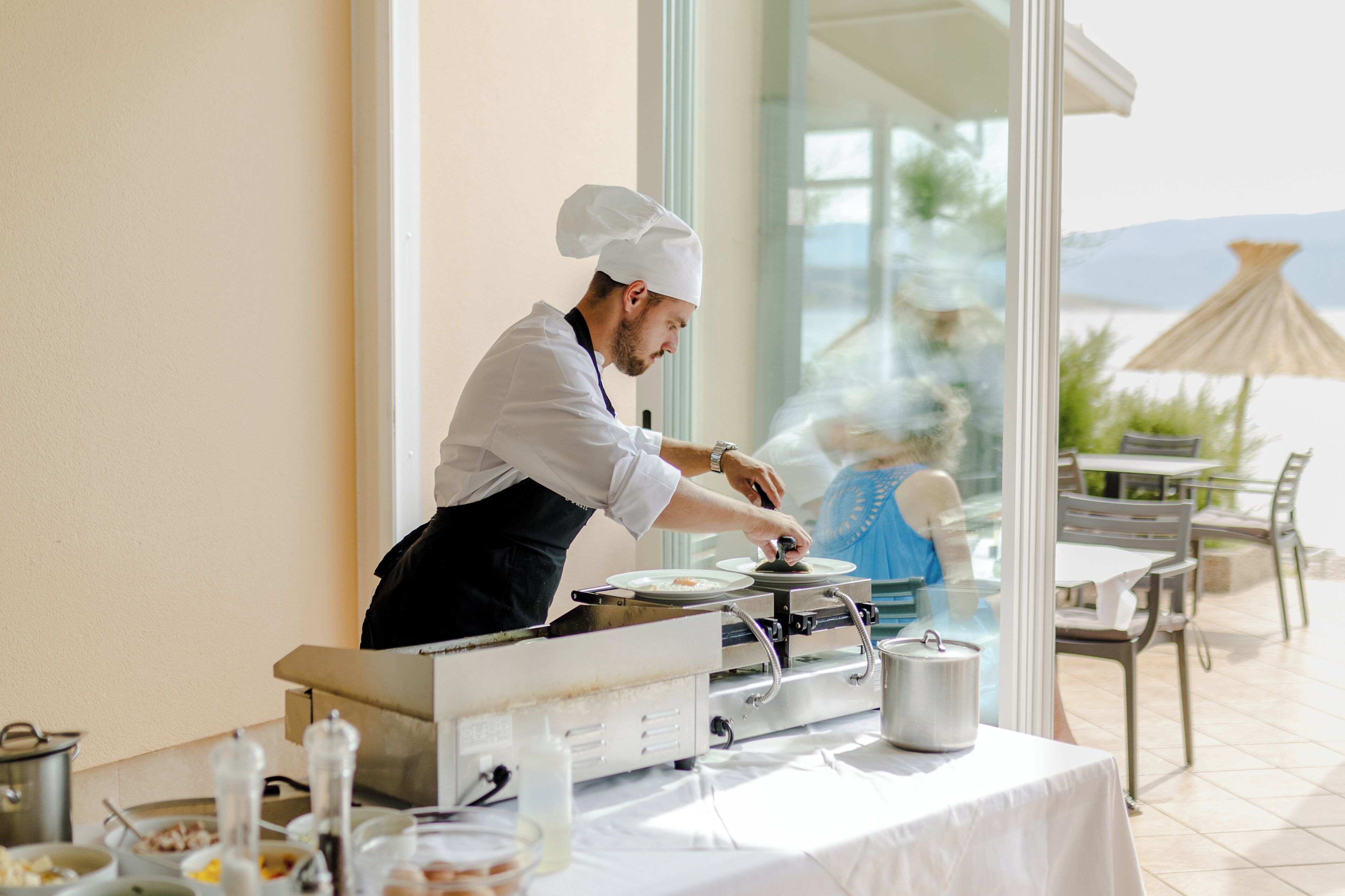 Um chef preparando comida em área de refeições ao ar livre, com vista para o mar e montanhas.