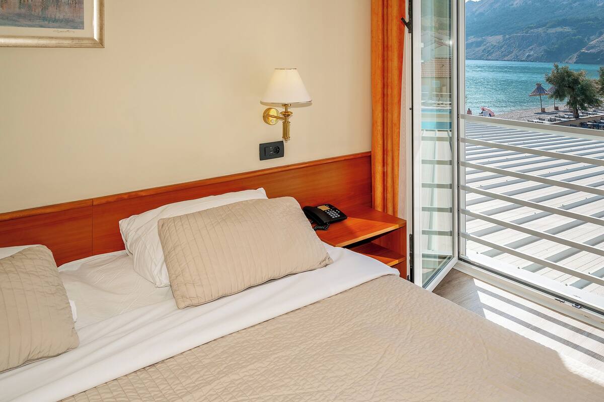 A hotel room with a bed, a bedside lamp, a phone, and a view of a beach through a large window.