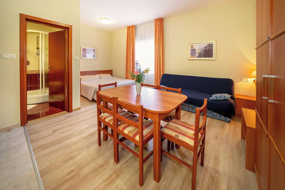 A hotel room with a wooden dining table, four chairs, a sofa, a bed, and a bathroom visible through an open door.