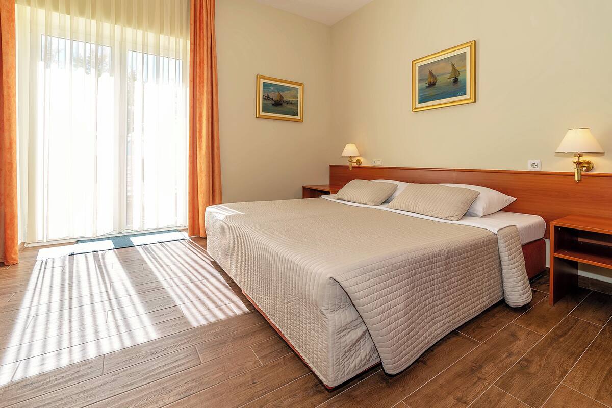 A hotel room with a large bed, wooden headboard, two bedside tables with lamps, a window with curtains, and two paintings on the walls.