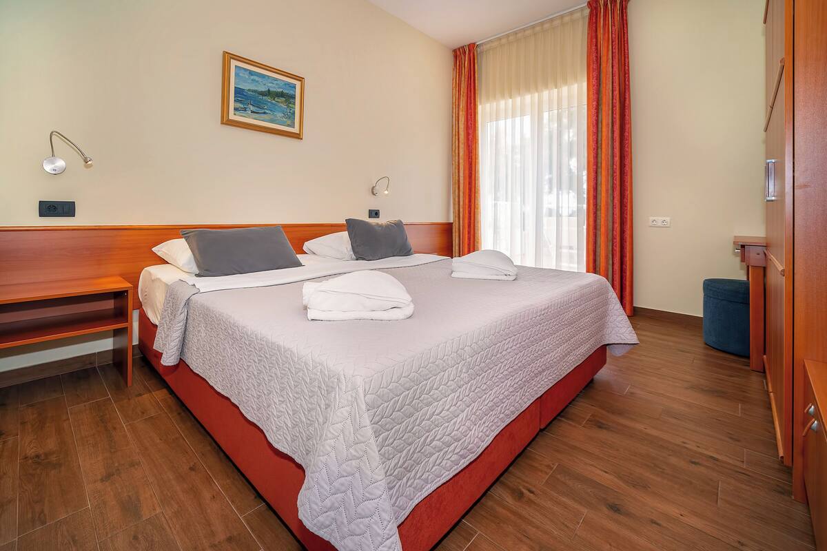 A hotel room with a large bed, two bedside tables, a bench, a painting on the wall, and a window with curtains.